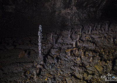 The image depicts a dark Lava cave interior featuring various rock formations. In the center, a tall, slender stalagmite stands prominently, surrounded by smaller, irregularly shaped formations. The cave floor is uneven and textured, displaying a mix of smooth and rugged surfaces, with shades of black and deep gray dominating the scene. Low lighting casts shadows and highlights the unique shapes of the stalagmites, creating an intriguing atmosphere.