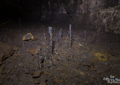 The image depicts an underground Lava cave floor characterized by dark, moist earth with various formations. Among the surface, numerous small, stalagmite-like structures rise, made of a shiny, dark material. The background reveals the cave walls, which appear rugged and shadowy, contributing to the overall dim ambiance of the scene. A hint of yellow tape is visible, suggesting some level of human exploration or marking within the cave environment.