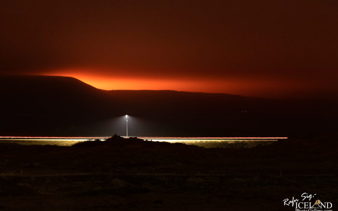 A nighttime landscape featuring a distant mountain range silhouetted against a vibrant orange and purple sky. A solitary lamppost stands illuminated in the foreground, casting light onto the ground, while faint streaks of light suggest the presence of moving vehicles on a road. The overall atmosphere is serene, with a sense of depth and tranquility emanating from the scene.