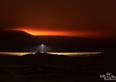 A nighttime landscape featuring a distant mountain range silhouetted against a vibrant orange and purple sky. A solitary lamppost stands illuminated in the foreground, casting light onto the ground, while faint streaks of light suggest the presence of moving vehicles on a road. The overall atmosphere is serene, with a sense of depth and tranquility emanating from the scene.