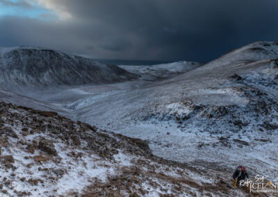 A rugged, snow-covered mountainous landscape under a dramatic sky. Sharp rocky outcrops and a softly rolling terrain stretch into the distance, with dark clouds looming overhead. A lone hiker is visible in the foreground, navigating the rocky slope, adding a sense of scale to the expansive wilderness.