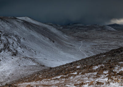 A vast, rugged landscape featuring snow-covered hills and rocky terrain under a dark, moody sky. In the distance, a winding path leads through the barren landscape, with small groups of hikers visible along the trail. The foreground displays a rocky slope with scattered stones, while the background showcases a range of hills fading into the clouds.