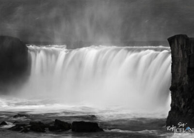 A black and white photograph of a cascading waterfall, where water flows dramatically over a cliff edge. Mist rises from where the water meets the river below, creating a sense of movement and tranquility. The rocky terrain surrounding the waterfall adds texture and contrast to the scene.