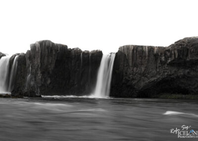 A serene landscape featuring a waterfall cascading over dark, rugged cliffs, surrounded by flowing water. The upper part of the image has a bright, nearly white sky, creating a contrast with the dark rock formations and the soft, blurred motion of the water. The overall atmosphere conveys a sense of tranquility and natural beauty.