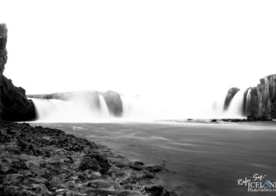 A monochrome photograph of a waterfall surrounded by rocky terrain. The water cascades down from a height, creating a misty effect as it flows into a river. The scene is mostly subdued in shades of gray, with soft, flowing water contrasting against the rugged rocks on the bank. The sky appears bright and almost washed out, emphasizing the tranquil, ethereal quality of the landscape.