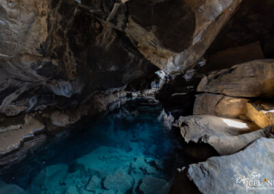 A dark, cavernous space with large, rugged rock formations framing a clear, turquoise pool of water. Light filters in from openings above, creating a serene atmosphere. Shadows play across the rocks and water, revealing textures and reflections. In the background, a few indistinct figures can be seen near the water's edge, adding a sense of scale to the scene.