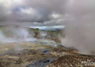 A landscape featuring a geothermal area with steam rising from the ground, set against a backdrop of rolling hills and overcast skies. The ground is rocky and uneven, with patches of green vegetation visible, and distant mountains are seen under a cloudy sky. The scene conveys a sense of natural geothermal activity and rugged terrain.