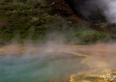 A geothermal hot spring with vibrant turquoise water surrounded by colorful mineral deposits in shades of yellow and orange. Steam rises from the surface, creating a misty atmosphere, while lush, green grass and rocky terrain are visible in the background.