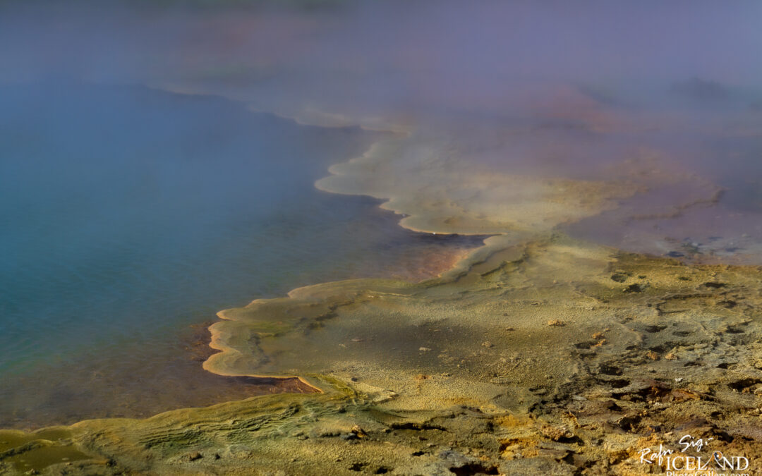 A close-up view of a geothermal area featuring vibrant, colorful mineral deposits along the edge of a steaming hot spring. The surface shows shades of blue and green water, with distinct layers of yellow and orange minerals creating a textured shoreline. Surrounding the area is a wispy fog that adds a mystical feel to the landscape, highlighting the natural beauty and unique geological features.