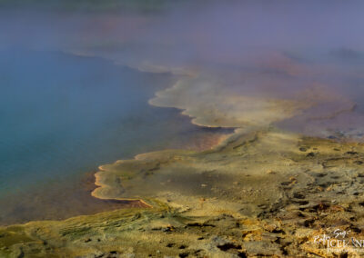 A close-up view of a geothermal area featuring vibrant, colorful mineral deposits along the edge of a steaming hot spring. The surface shows shades of blue and green water, with distinct layers of yellow and orange minerals creating a textured shoreline. Surrounding the area is a wispy fog that adds a mystical feel to the landscape, highlighting the natural beauty and unique geological features.
