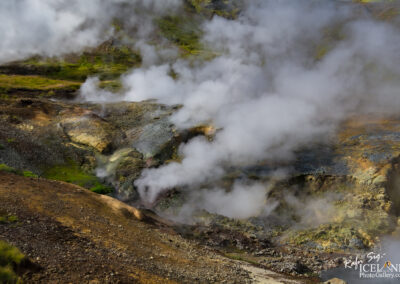 A geothermal landscape features steaming vents and colorful mineral deposits, surrounded by rocky terrain and patches of green vegetation. Thick white clouds of steam rise from the ground, creating a mystical atmosphere. The region displays a mix of earth tones, including browns, greens, and hints of blue, with small mounds and ridges reflecting the geothermal activity.