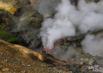 A geothermal area with steam rising from the ground, surrounded by a rugged landscape featuring earthy tones and patches of green moss. The colors range from yellow to dark browns, highlighting the mineral-rich terrain. Steam clouds billow upwards, creating a mystical atmosphere in the geothermal landscape.