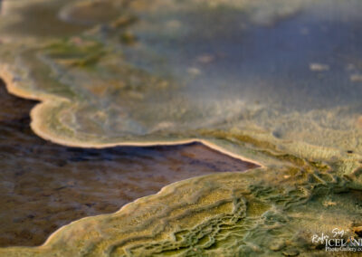 A close-up view of colorful, layered mineral formations around a hot spring, featuring shades of green, brown, and a hint of blue in the background water. The texture of the minerals is intricate, creating an organic pattern that showcases the natural geothermal activity in the area.