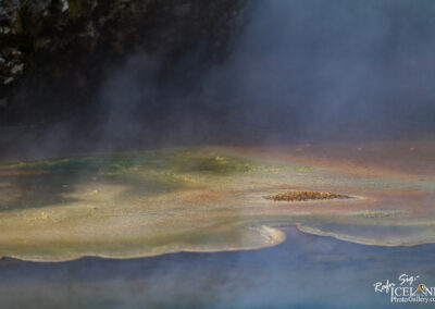A close-up view of a geothermal feature, showcasing a colorful, textured surface with shades of yellow, green, and blue. Steam rises from the edges, creating a misty atmosphere. The ground appears uneven with a central mound, typical of hot spring environments. Natural rock formations are partially visible in the background.