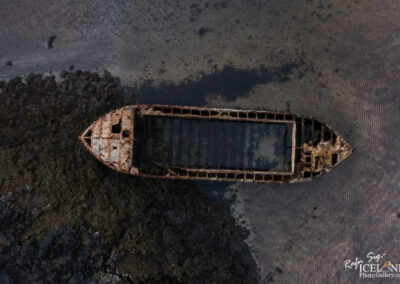 An aerial view of a rusted shipwreck sitting in shallow waters, surrounded by patches of dark seaweed and a sandy seabed. The ship is partially submerged, showcasing its decayed structure, with visible openings and a weathered bow. The surrounding landscape features rippled patterns in the sand, blending into the serene water.