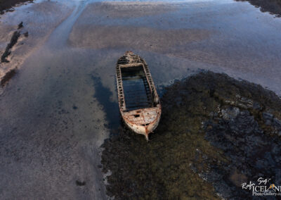 An aerial view of an old, abandoned boat resting on a shallow tidal flat, surrounded by seaweed and rocky terrain. The boat shows signs of rust and decay, with its hull partially submerged in clear, shallow water that reflects the sky. The surrounding area consists of smooth sand patterns and patches of marine vegetation.