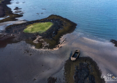 An aerial view of a coastal landscape featuring a shipwreck on the shore. The wreck, partially submerged in shallow water, is surrounded by dark rocks and patches of sand, while a small grassy island is visible nearby. The calm blue waters extend in the background, with gentle waves lapping at the shoreline. The scene conveys a sense of tranquility and natural beauty.
