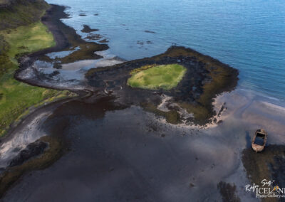 An aerial view of a coastal landscape featuring a mix of lush green grass, dark rocky areas, and a calm blue sea. A small grassy island is visible in the water, surrounded by shallow, dark sediment. In the foreground, a dilapidated boat rests partially on the shore, adding a rustic element to the natural scenery. The overall atmosphere is serene and reflects the beauty of an untouched coastal environment.