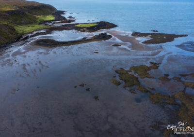 A panoramic view of a coastal landscape featuring a serene shoreline with a mix of wetland areas and rocky outcrops. The foreground shows shallow waters reflecting the sky and revealing patches of sand and seaweed. In the background, lush green vegetation contrasts with the darker rocky cliffs that rise toward the horizon. The calm ocean extends into the distance under a clear blue sky.