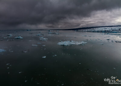 A serene, icy landscape features floating icebergs in a dark, still body of water, under a moody, overcast sky. The distant shoreline is lined with a glacier, contributing to the dramatic atmosphere of the scene. The colors are predominantly cool, with shades of blue and gray, creating a tranquil yet hauntingly beautiful vista.