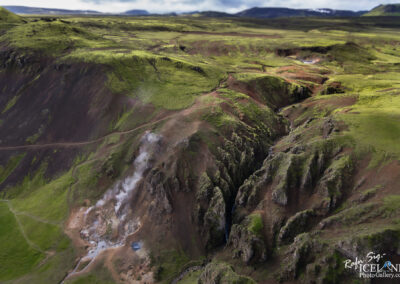 Aerial view of a rugged landscape featuring green hills and steep cliffs, interspersed with volcanic rock formations. Steam rises from geothermal activity in the lower left corner, while a small stream winds through the crevices. The sky is partially cloudy, providing a dramatic backdrop to the natural scenery.