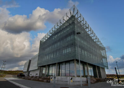 A modern, glass-fronted building with a unique architectural design, featuring angular shapes and a transparent facade. The structure has multiple levels and is surrounded by a paved area with parking spaces. In the foreground, there are large stones and lampposts lining the walkway. The sky is partly cloudy, with blue and grey hues.