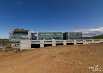 A modern building with large glass windows and a distinctive angular design sits atop a concrete foundation. The structure features multiple levels and is surrounded by a gravel and dirt landscape, with a few parked cars visible in front. The sky above is clear and blue, while green hills are in the background, suggesting a natural setting.