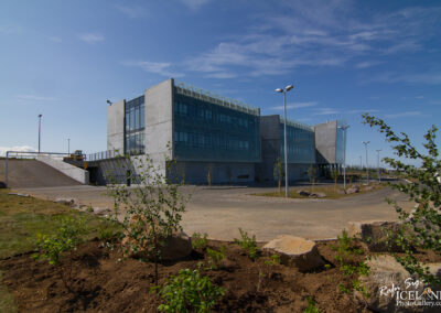 A modern, angular concrete building is depicted against a clear blue sky. The structure features large glass windows and a unique pattern on the upper section. In the foreground, small green shrubs are planted, alongside a few rocks. A sloped driveway leads up to the entrance, with streetlights scattered around the area. The scene is well-lit, suggesting a sunny day.