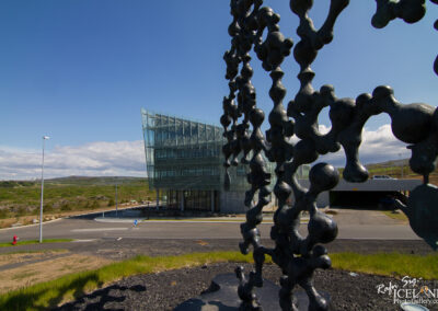 A modern glass building with a unique angular design stands in the background, surrounded by green hillside and under a clear blue sky. In the foreground, a large abstract sculpture with a cluster of rounded, connected shapes is prominently displayed. A paved road curves around the scene, and a streetlight is visible along the edge. The area appears to be well-maintained with patches of grass and a hint of urban development in the distant landscape.