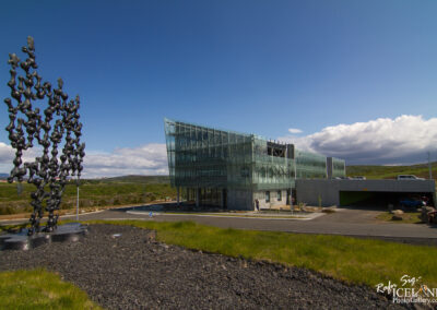A modern glass building with an angular design is situated amidst green landscape and blue skies. In the foreground, there is a large, abstract black sculpture made of numerous interconnected figures. The building features large, transparent windows and a concrete base, with some people visible near the entrance. Surrounding the structure are patches of grass and a circular driveway, with a few vehicles parked nearby.