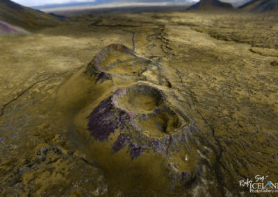 Aerial view of a rocky, barren landscape featuring several small volcanic craters. The ground is predominantly yellowish with patches of dark purple and brown, suggesting a rugged and uneven terrain. In the background, a distant horizon with muted colors presents a sense of vastness and isolation. The sky is partly cloudy, enhancing the stark beauty of the volcanic environment.