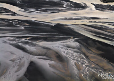 An aerial view of a river delta, displaying intricate patterns of black sand and flowing water. The image features a mix of dark and light tones, with winding waterways and smooth curves creating a striking contrast against the textured landscape. The overall composition highlights the natural beauty and complexity of the terrain.