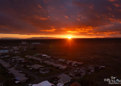 A vibrant sunset over a sparse landscape, with the sun dipping below the horizon, casting a warm orange glow across the sky. In the foreground, a residential area with rooftops and scattered vehicles is visible, while a winding road leads to commercial buildings in the distance. The mountains are silhouetted against the colorful sky, creating a picturesque scene of tranquility and natural beauty.