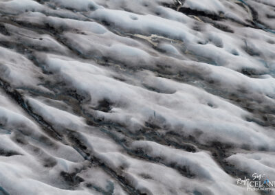 A close-up view of a glacier surface, featuring a mix of smooth, white ice and darker, textured areas, indicating underlying rock or debris. The scene is marked by undulating patterns and varying shades of blue and gray, showcasing the intricate details of melting ice and the natural formations created by the glacial environment.