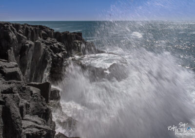 A dramatic seascape featuring rugged black volcanic cliffs adjacent to the ocean. The waves crash forcefully against the rocks, creating a mist of water droplets that glisten in the sunlight. The sky is clear with a few wisps of clouds, and the shimmering sea stretches toward the horizon.