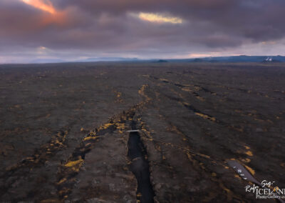 Aerial view of a vast, desolate landscape with black lava fields stretching to the horizon, interspersed with patches of yellowish vegetation. A winding stream carved through the terrain reflects the cloudy sky above, which is filled with shades of gray and hints of orange from the sunset. A small wooden bridge crosses the stream, and a few vehicles are parked nearby, adding a touch of scale to the expansive scene. Silhouetted mountains can be seen in the distance, partially shrouded in mist.