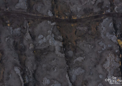 An aerial view of a rugged volcanic landscape featuring black volcanic rock formations and a narrow dirt path weaving through the terrain. The surface shows a mix of dark, rough textures interspersed with patches of lighter-colored rock and some sparse greenery, suggesting a harsh but striking environment shaped by volcanic activity.