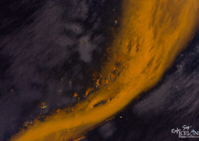 An aerial view of a dark landscape with a prominent, swirling band of bright orange river. The contrast between the vibrant orange and the deep black and gray tones creates a striking visual effect, reminiscent of abstract art. The surface displays varying textures and subtle details, suggesting natural elements.