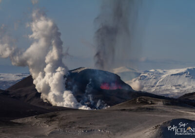 A dramatic landscape showcasing an erupting volcano. Thick white plumes of smoke rise from the volcano's crater, while dark ash clouds billow into the sky. In the foreground, the volcanic terrain is rugged and gray, with scattered patches of snow in the background. The distant mountains are partially covered in snow, contrasting with the volcanic activity in the foreground. The scene captures a blend of natural beauty and raw geological power.