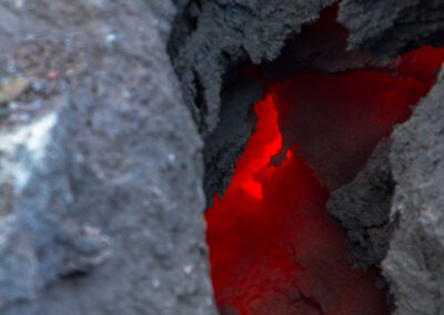 A close-up view of a crack in volcanic rock, revealing glowing red lava beneath the surface. The texture of the dark, rough rock contrasts with the bright, intense color of the molten lava seen shining through the fissure.