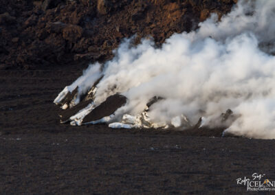 A close-up view of volcanic activity, featuring dark, rocky terrain with steam billowing from the surface. The steam rises from areas where molten lava meets cooler surfaces, creating a contrast between the dark volcanic rock and the white, steamy vapor. The surrounding environment appears rugged and barren, indicating the dynamic and raw nature of the landscape.