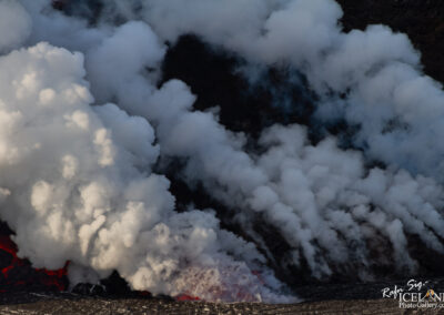 A dramatic view of smoke and steam rising from volcanic activity. The foreground features billowing white clouds of steam, while hints of glowing red lava can be seen at the base, contrasting against the dark, rocky terrain. The scene captures the raw power and beauty of nature in a volcanic landscape.