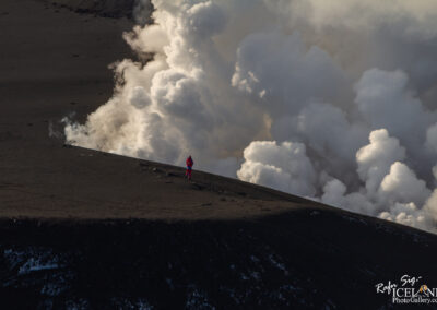 A person in a bright red outfit stands on the edge of a volcanic landscape, surrounded by billowing white steam and clouds. The ground is dark and textured, indicating volcanic rock, while the background features more steam rising from the volcanic activity. The scene captures a sense of isolation and the dramatic interplay between the human figure and the natural environment.