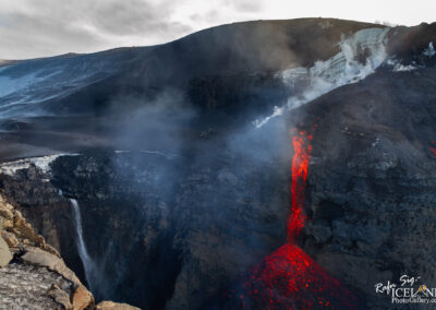 A dramatic landscape featuring an active volcano with flowing lava cascading down a rocky cliff. Smoke and steam rise into the air, contrasting with the dark volcanic rock and the lighter icy surfaces above. A waterfall can be seen tumbling into the chasm below, adding to the rugged beauty of the scene. The sky is overcast, creating a somber atmosphere.