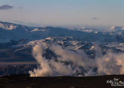 A panoramic view of a mountainous landscape blanketed in snow, with rugged peaks and ridges under a soft blue sky. Wisps of clouds drift through the valleys, and the sunlight casts a gentle glow on the terrain, creating a serene and picturesque scene of nature's beauty.