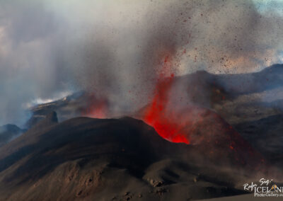A dramatic volcanic eruption scene showing a dark, rugged landscape with vibrant red lava spouting from the crater. Ash and smoke billow upwards into a partly cloudy sky, creating a contrasting backdrop to the fiery display. Scattered fragments of volcanic rock and ash are visible in the air, enhancing the intensity of the eruption.