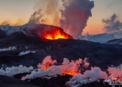 A dramatic landscape capturing an erupting volcano at sunset, with bright orange lava spilling from the crater and contrasting against the dark volcanic rock. Thick plumes of smoke and steam rise into the twilight sky, while the background features distant snowy mountains, creating a striking combination of elements in the scene. The vibrant colors of the molten lava and the soft hues of the sky add to the intensity of the moment.
