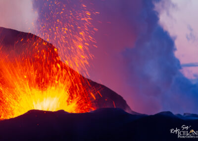 A vibrant eruption of a volcano, with bright orange and yellow lava bursting upwards against a backdrop of purple and dark clouds. The molten rock projects upwards, creating a dramatic and fiery display at the summit, while ash and smoke billow into the atmosphere. The surrounding landscape features dark volcanic soil.