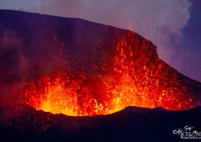 A close-up view of an erupting volcano, showcasing a dramatic display of bright orange and red lava spewing outwards against a dark, smoky backdrop. The glowing lava contrasts sharply with the surrounding landscape, capturing the raw power and intensity of the volcanic activity.