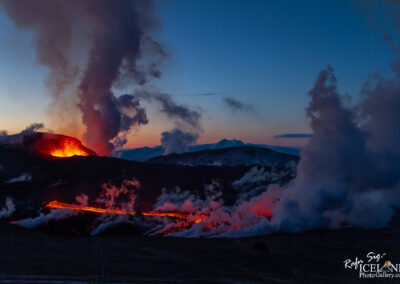 A dramatic volcanic landscape at dusk, featuring an erupting volcano with bright orange lava spewing from its crater. Thick plumes of steam and smoke rise into the twilight sky, contrasting with the dark silhouette of surrounding mountains. The ground is illuminated by the flowing lava, creating a vibrant river of red against the dusky background.